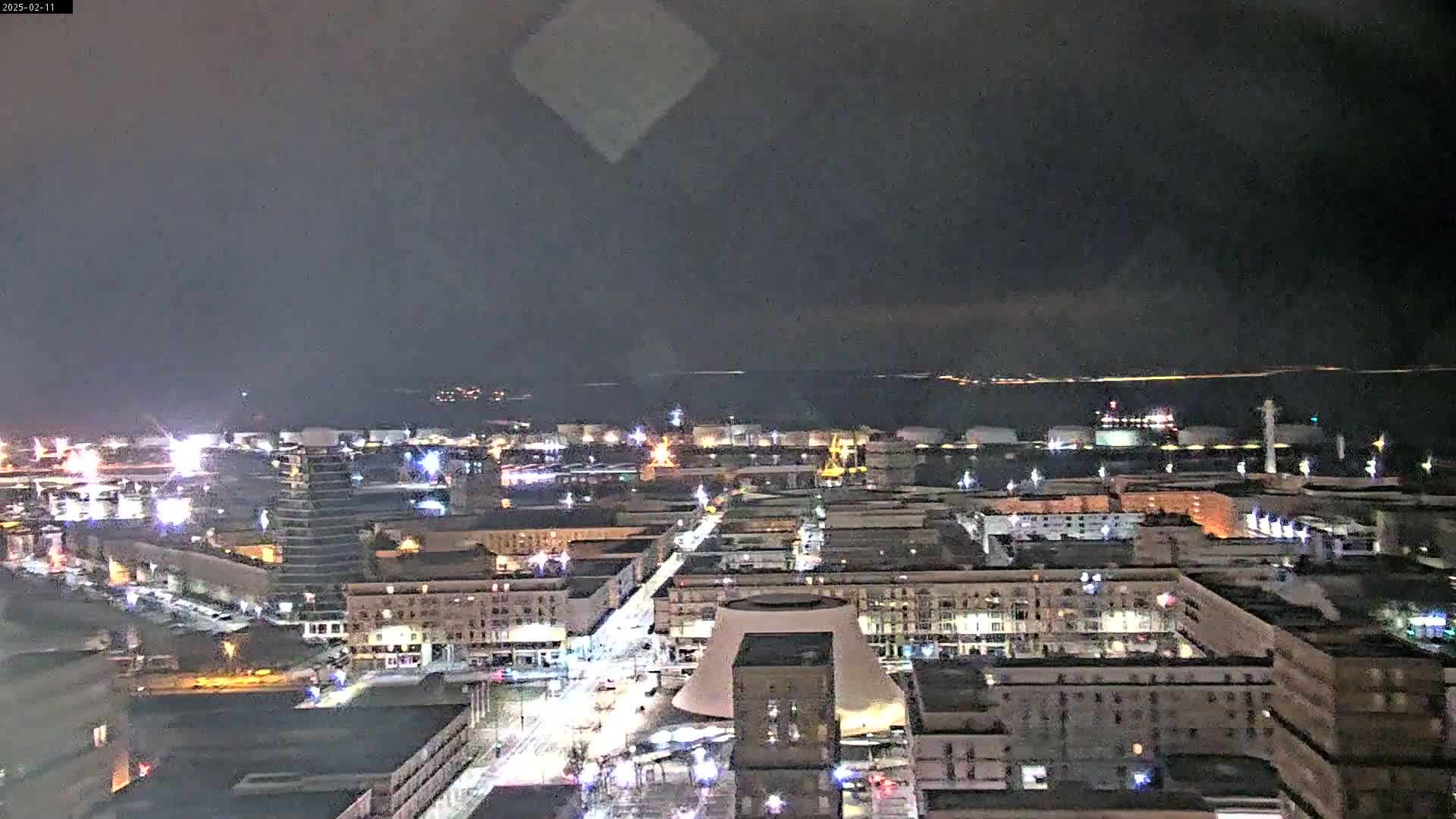 A nighttime aerial view of a city illuminated by streetlights and building lights, overlooking a dark body of water under a cloudy sky.