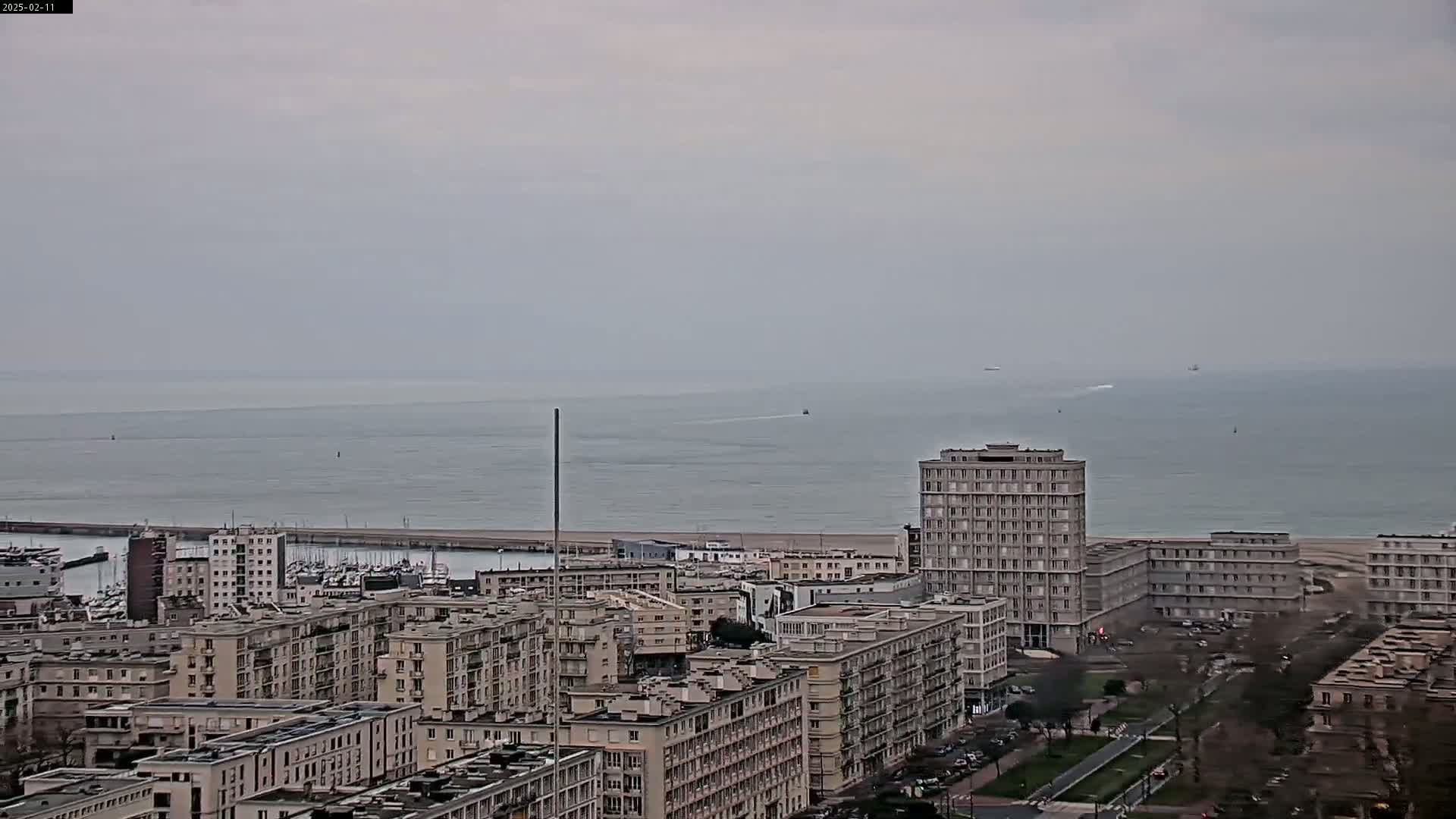 A coastal city with numerous apartment buildings is visible under an overcast sky, overlooking a calm sea with a few vessels in the distance.