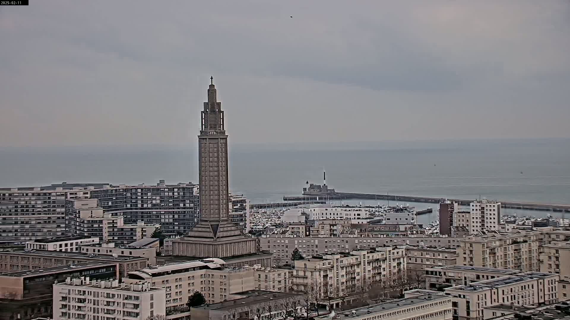 A tall, slender tower stands amidst a city overlooking a calm, gray sea under an overcast sky.