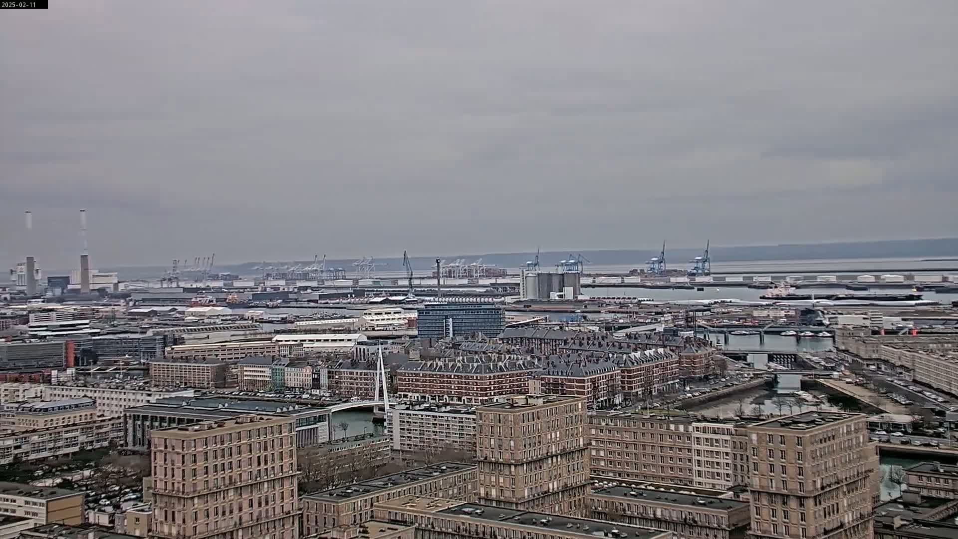 An overcast day reveals a sprawling cityscape featuring a port with numerous cranes, industrial structures, and residential buildings, all situated near a body of water.