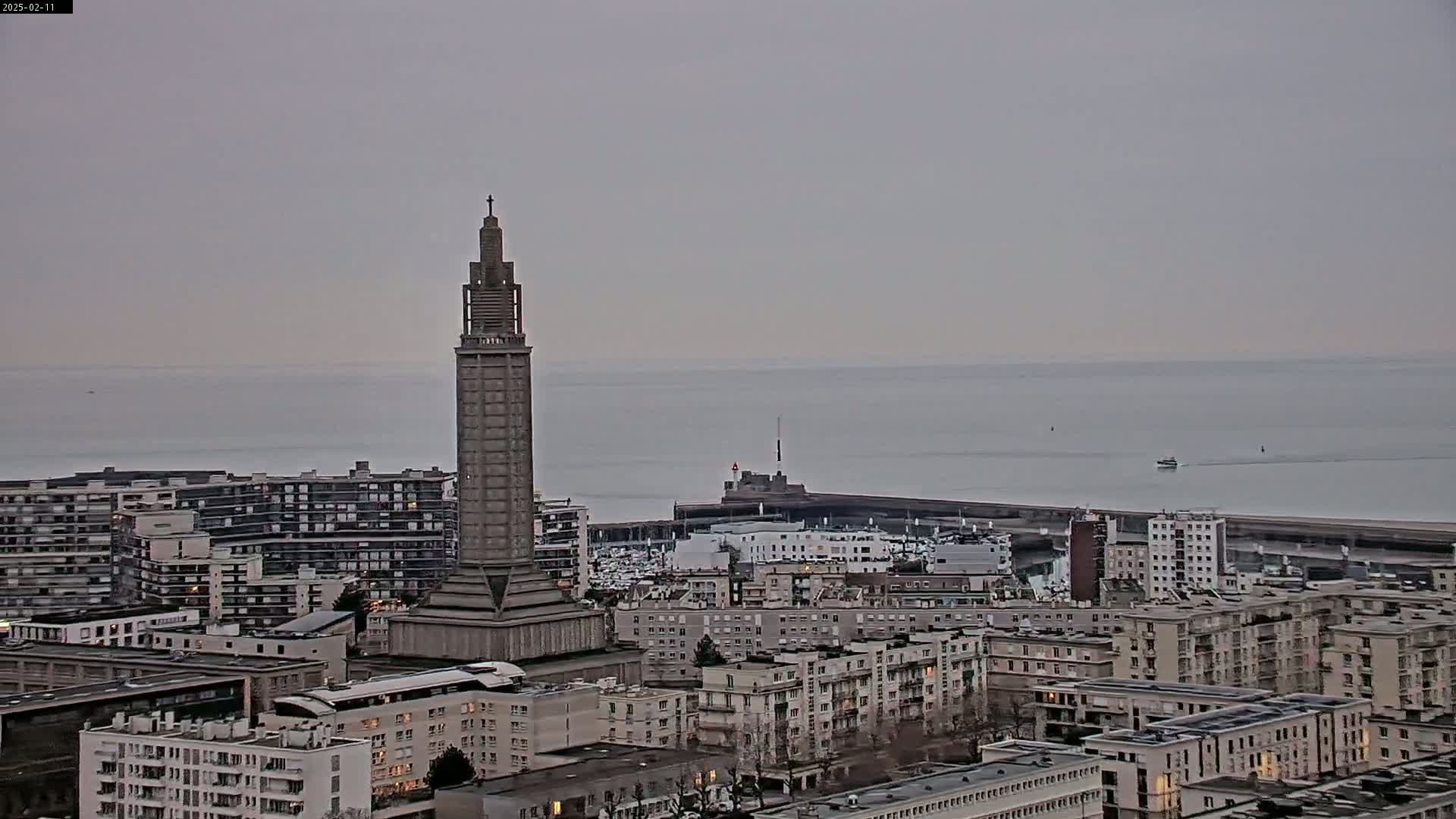A tall, slender tower stands prominently in a cityscape overlooking a calm, gray sea under an overcast sky.
