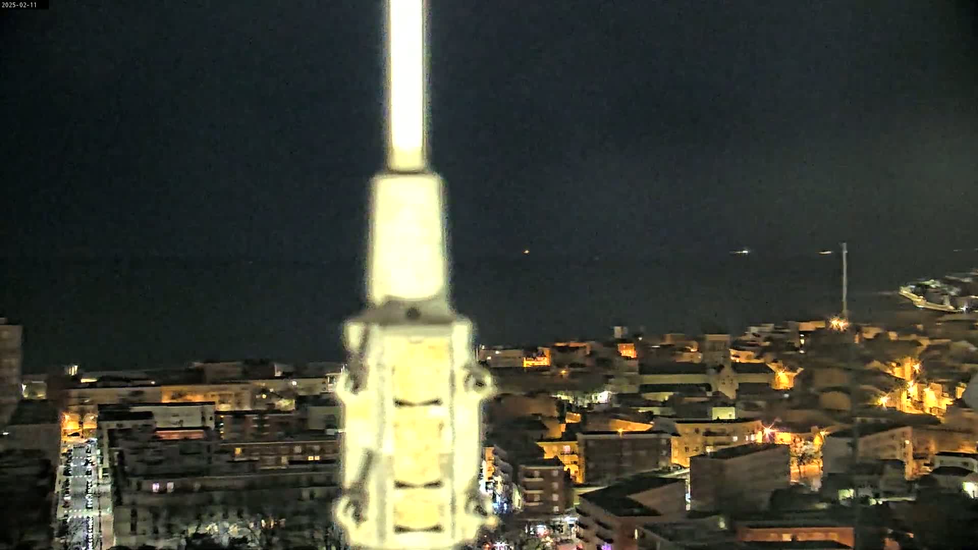 A nighttime view from a high vantage point shows a coastal town with illuminated buildings and a dark body of water beyond it, all seen through a tall, light-colored structure in the foreground.