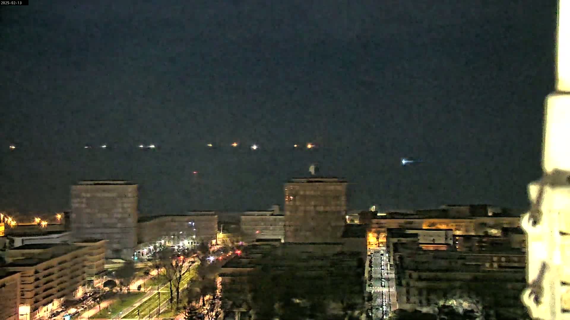 A nighttime view of a city skyline with several buildings illuminated, overlooking a body of water where distant lights are visible.