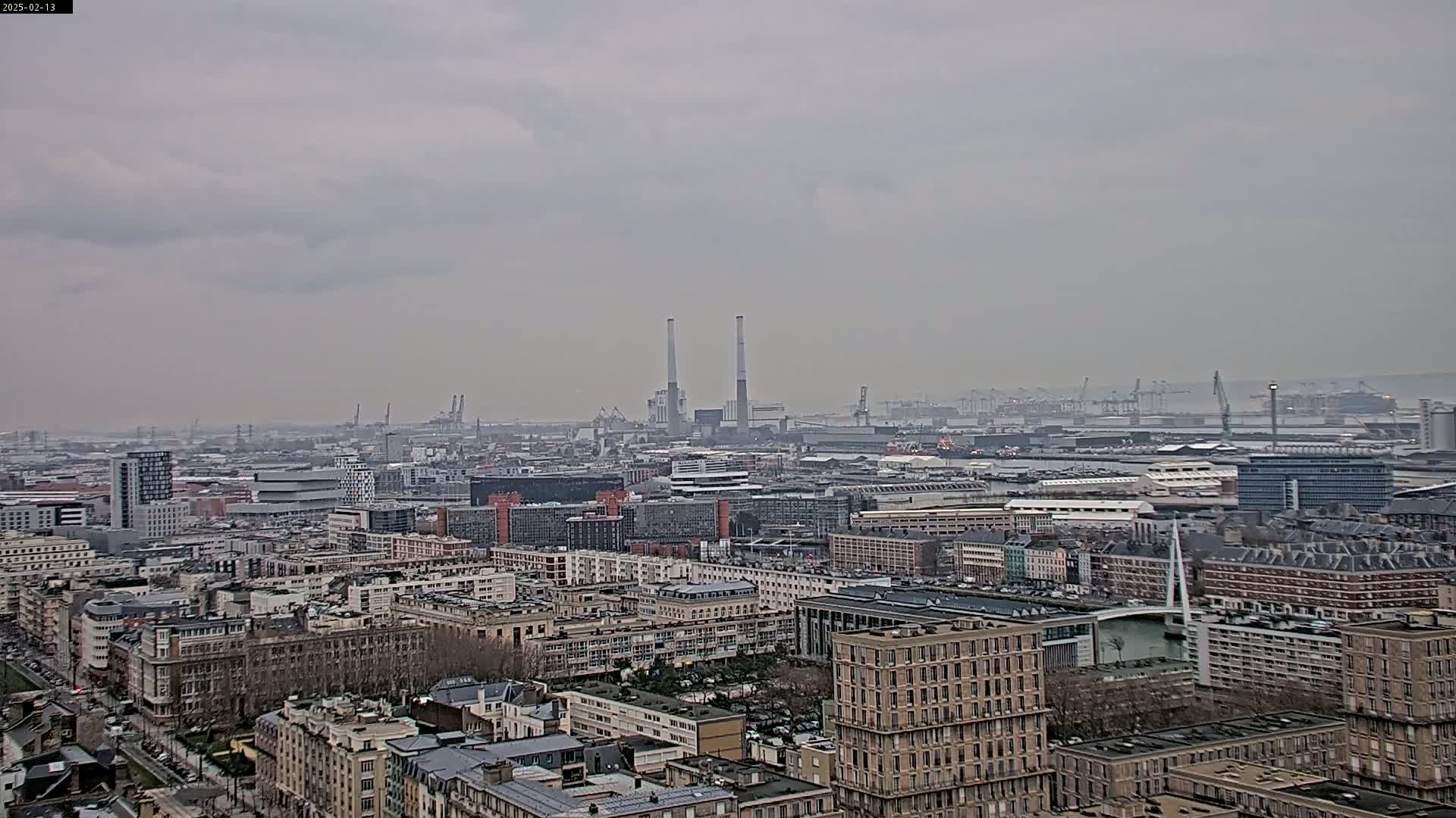 A hazy, overcast day reveals a sprawling cityscape, featuring numerous buildings, a port area with industrial structures and cranes in the distance, and two prominent smokestacks.