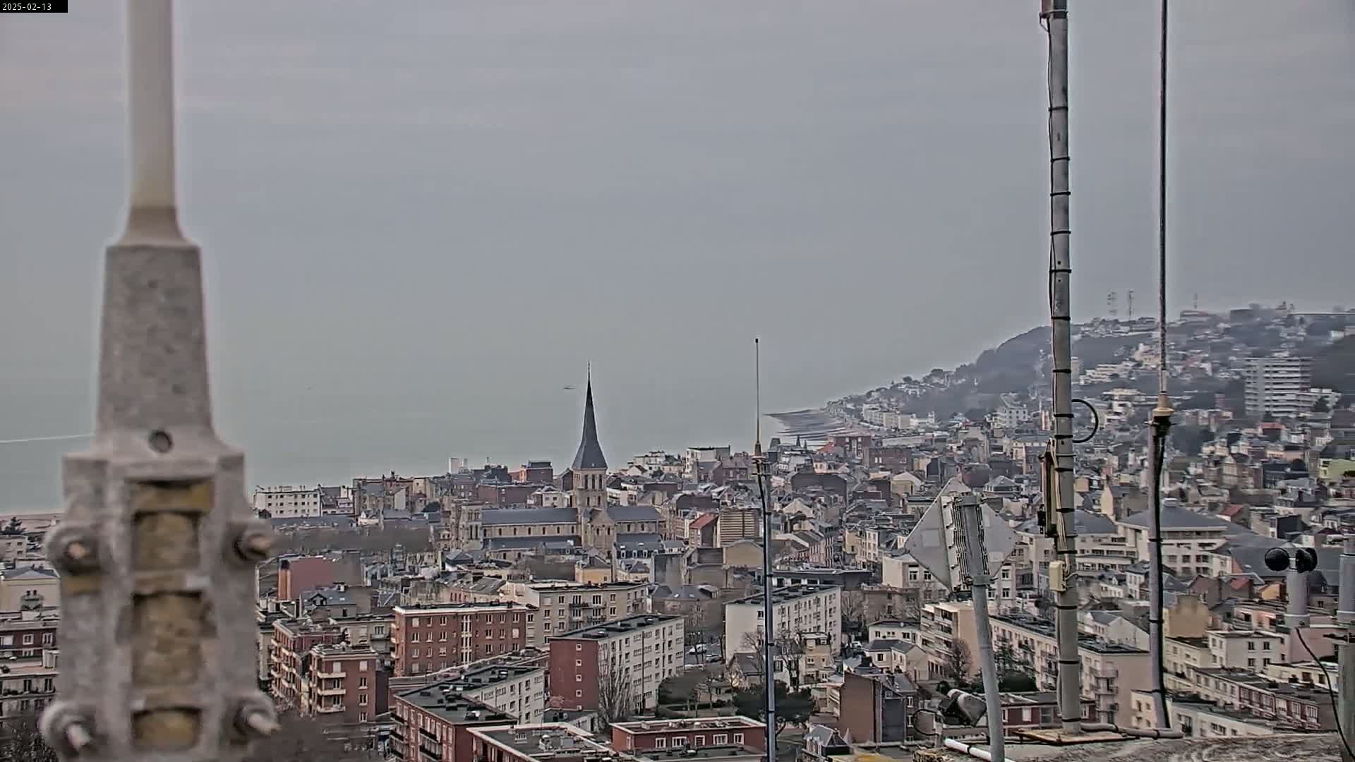 A high-angle, long shot on an overcast day shows a coastal city with a church steeple in the center, a body of water in the background, and several antenna towers in the foreground.