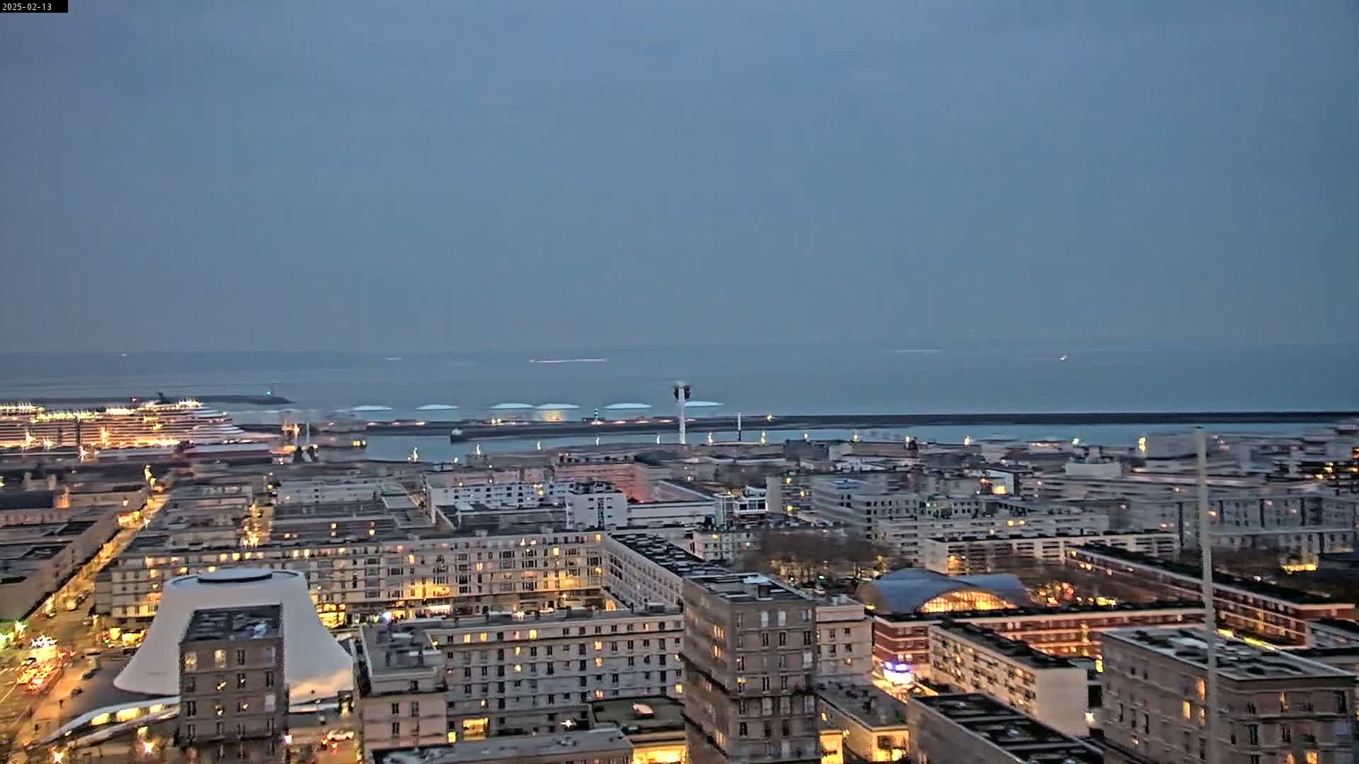 An aerial view at twilight shows a city with a harbor, cruise ships, and a calm sea in the distance.