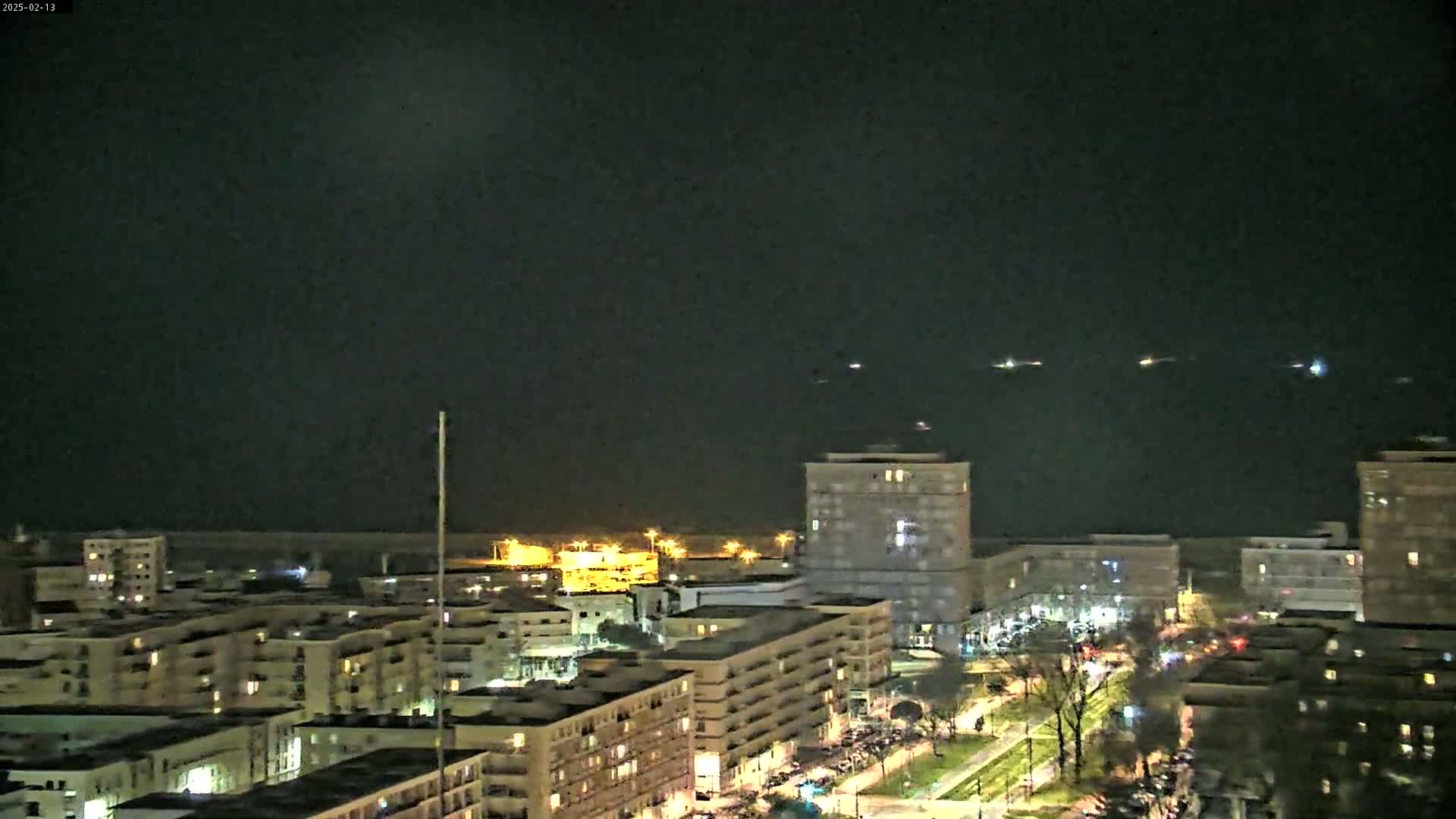 A nighttime view of a city near a body of water, showing several multi-story buildings with lights on, and a few distant lights visible on the water.