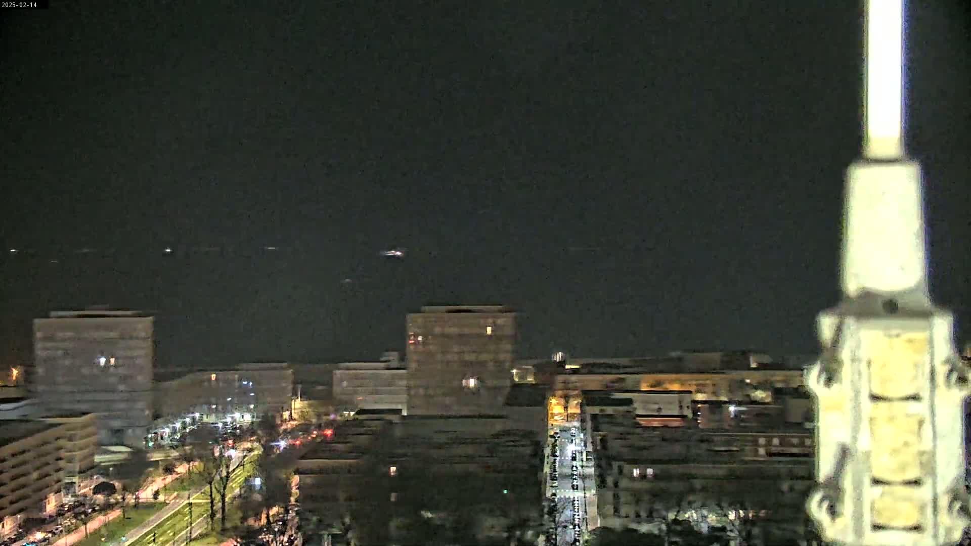 A nighttime, high-angle view shows city buildings and streets with some vehicle lights, a dark body of water in the distance, and a clear, dark sky.