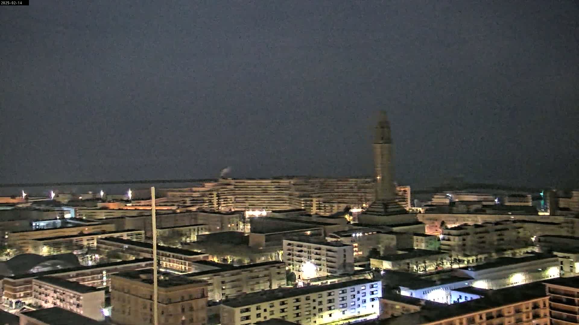 A nighttime cityscape with a tall tower is visible, under a dark, overcast sky.