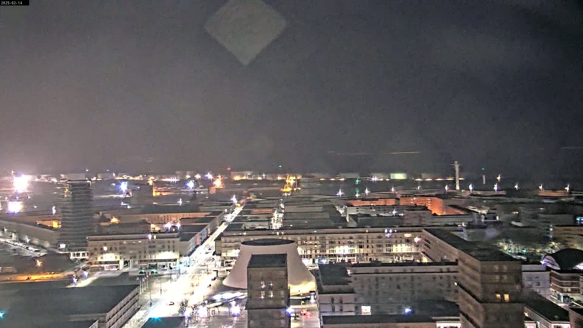 A nighttime, aerial view of a city illuminated by streetlights and distant lights, possibly on a body of water, under a dark, cloudy sky.