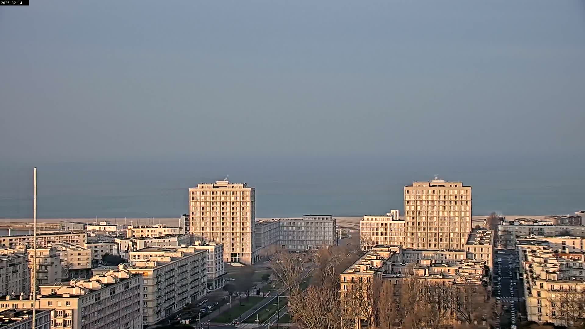 A coastal city with several high-rise buildings is seen under a hazy sky, with a calm sea in the background.
