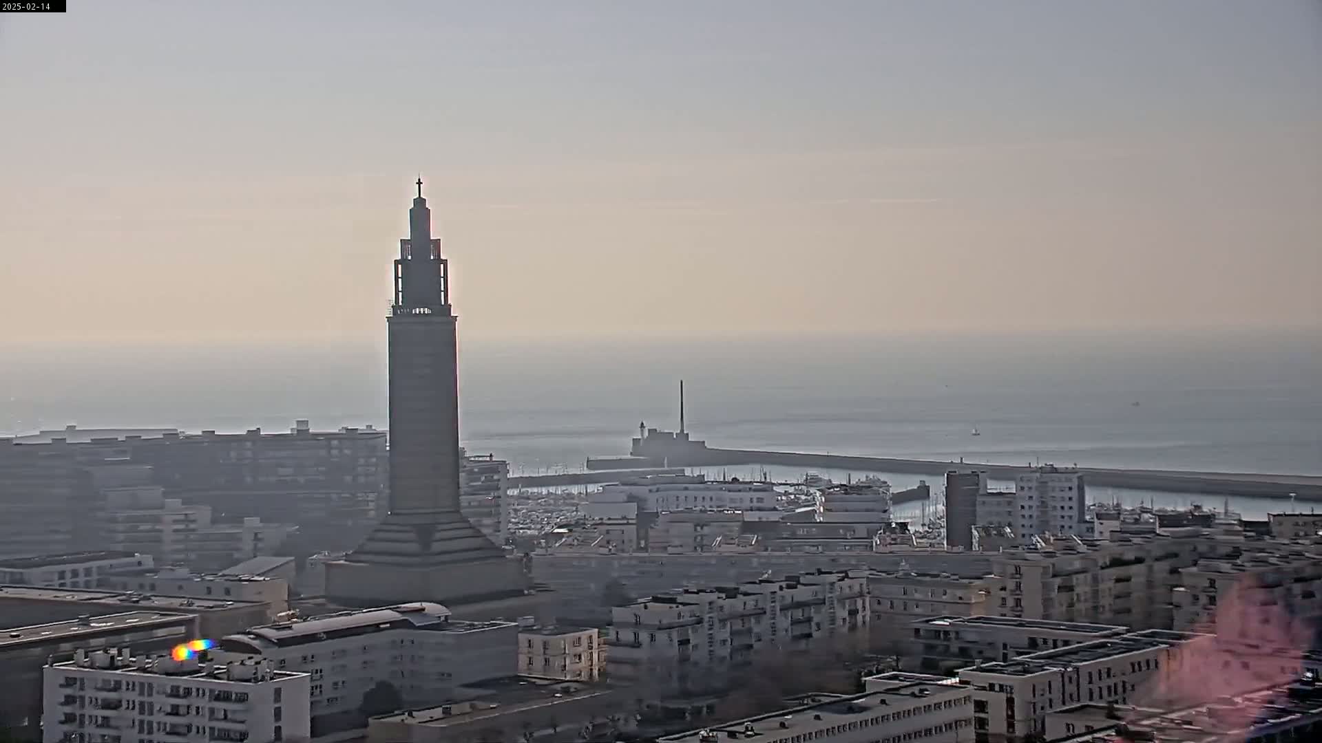 A tall, slender tower stands prominently in a city overlooking a hazy, calm ocean and a harbor filled with boats.