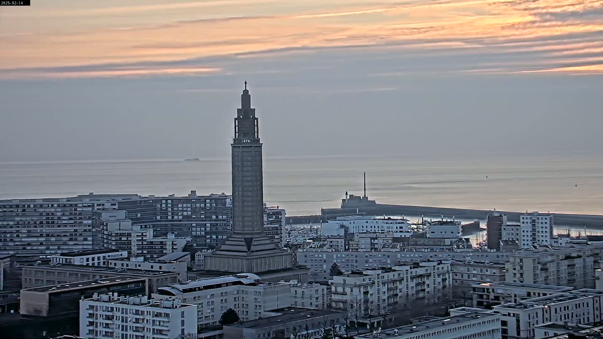 A tall, slender tower stands prominently in a city overlooking a calm sea under a pale, pinkish sunset sky.