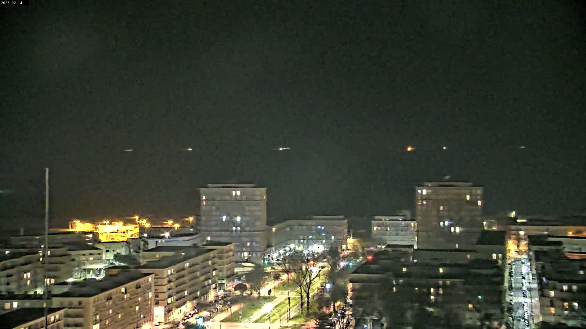 A nighttime view of a city skyline with several multi-story buildings illuminated, under a dark sky with a few distant lights possibly from ships or aircraft.