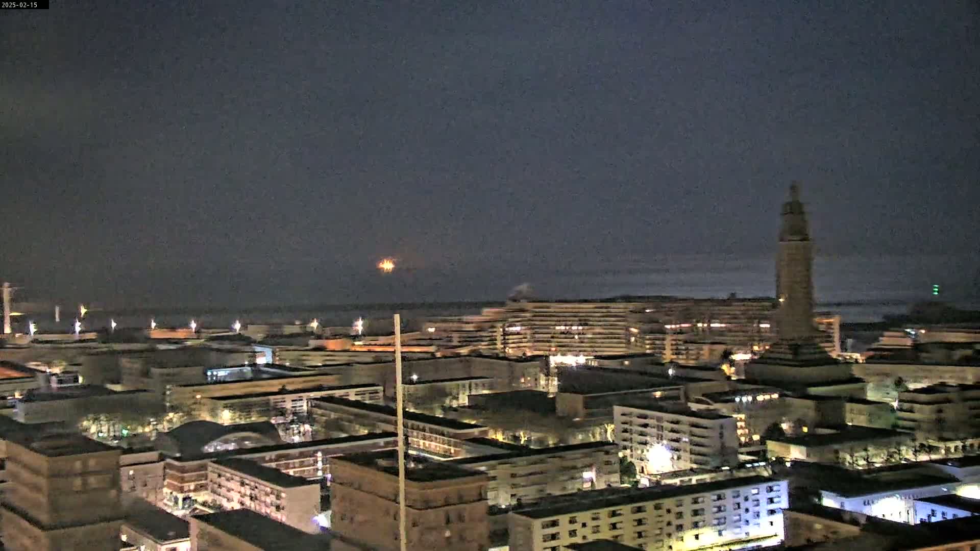 A nighttime aerial view of a city illuminated with lights, under a dark sky with a distant, faint light source possibly a flare or star.