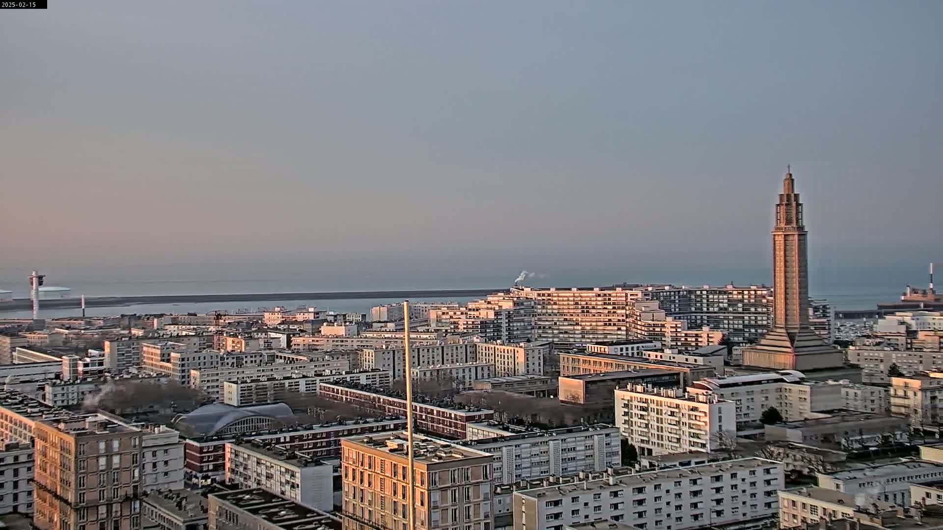 A coastal city with numerous multi-story apartment buildings under a hazy, pale sky, featuring a tall, slender tower in the center.