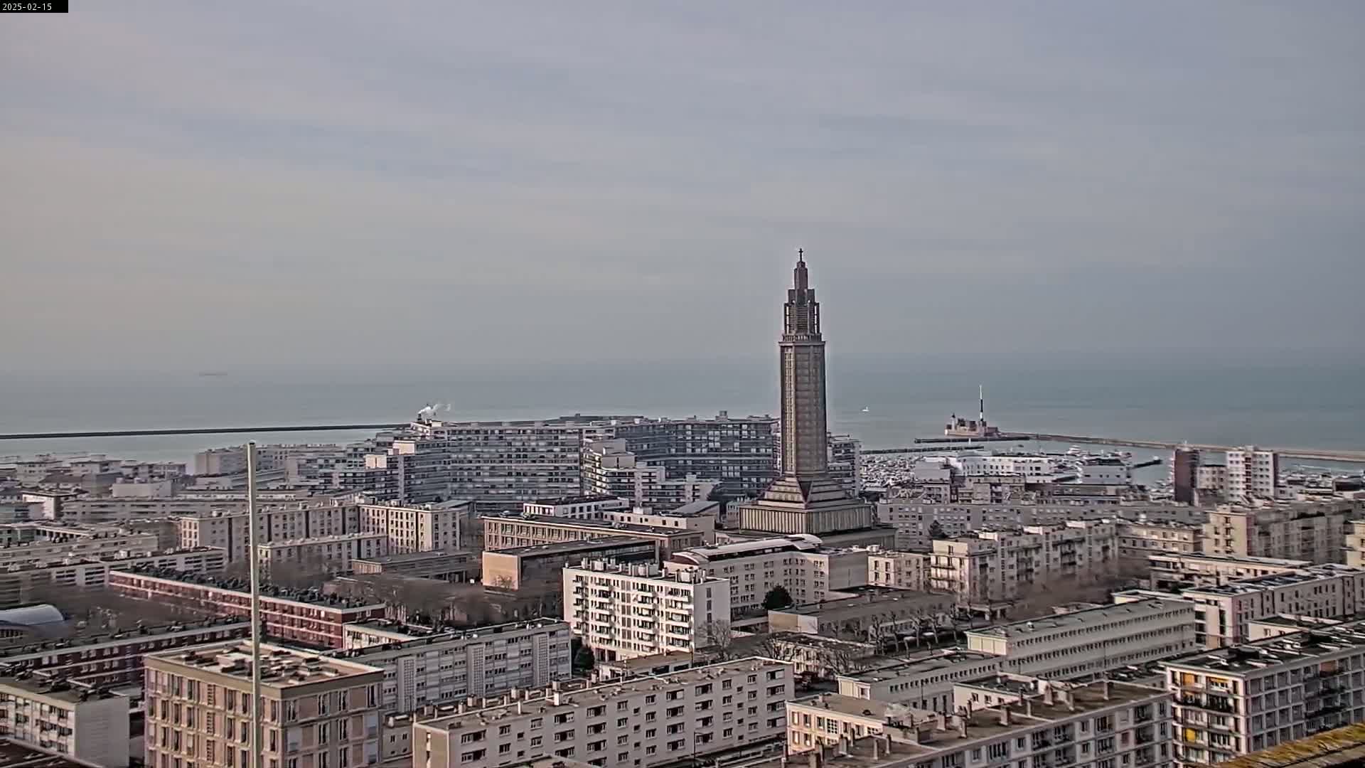 A tall, slender tower stands prominently in a cityscape overlooking a calm sea under a cloudy sky.
