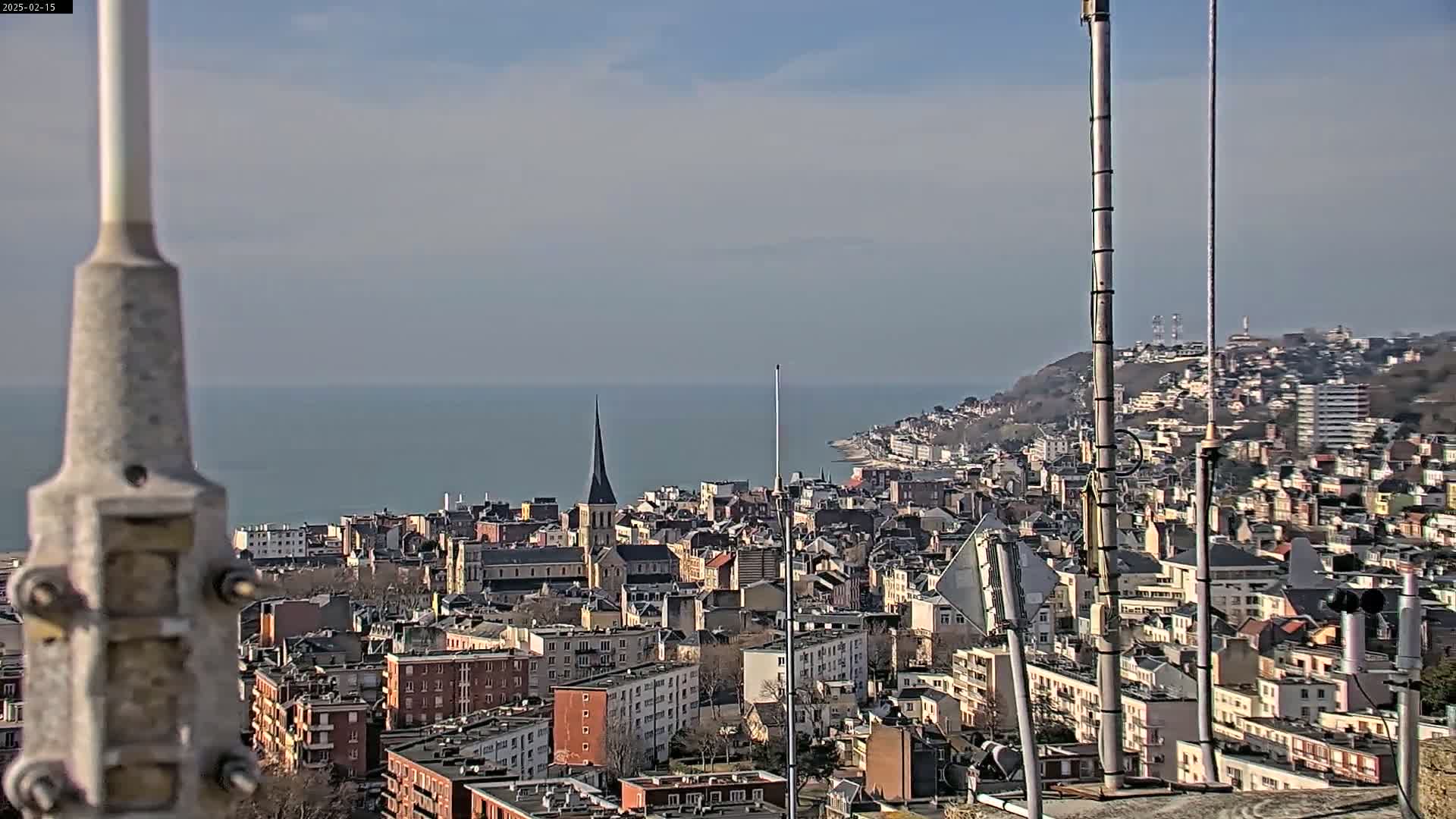 A coastal city with a church spire is visible under a clear, sunny sky, with a calm sea in the background.