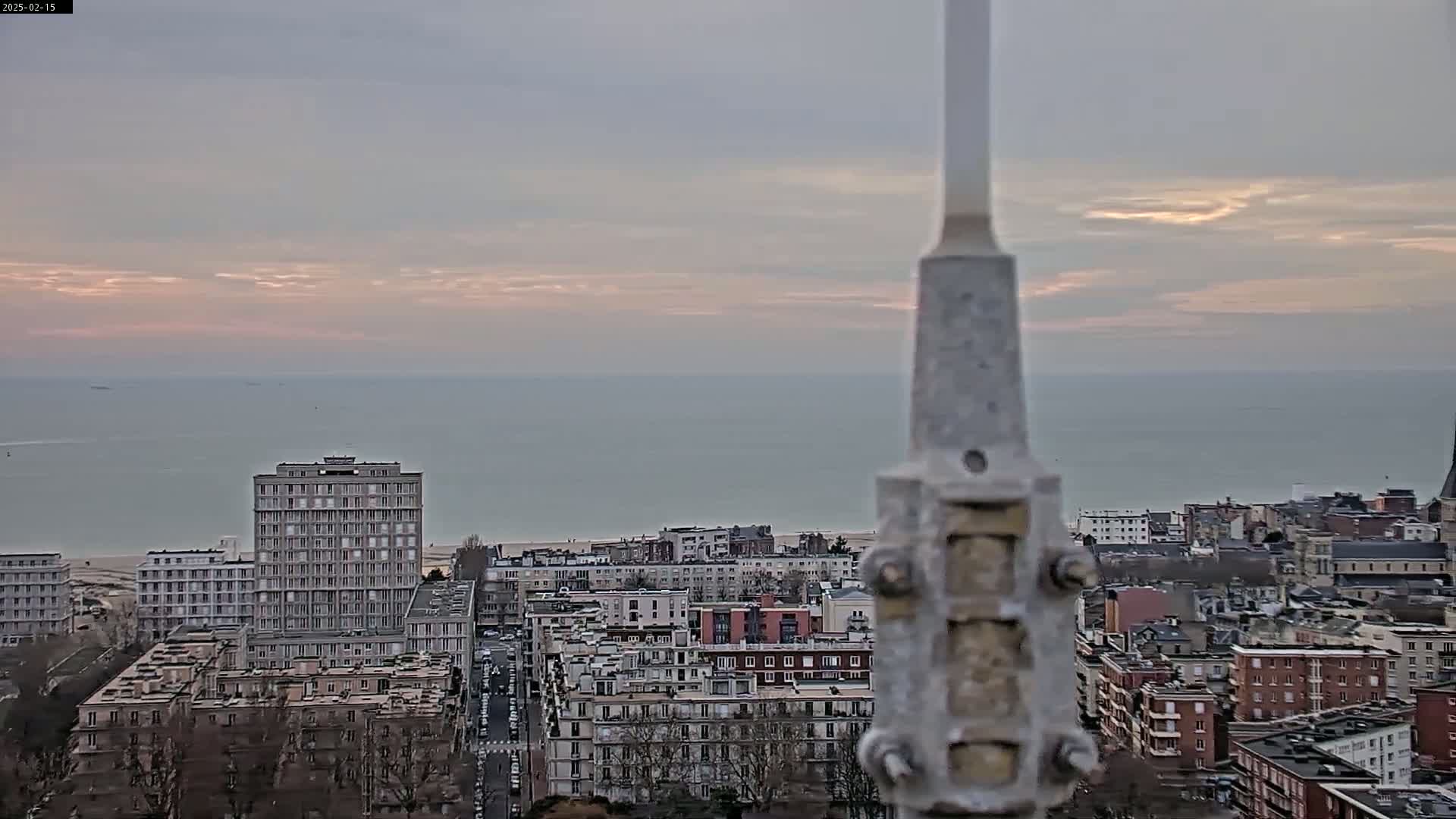 A coastal city scene at sunset under a cloudy sky, viewed from a height, showing a section of buildings and a calm sea in the distance.