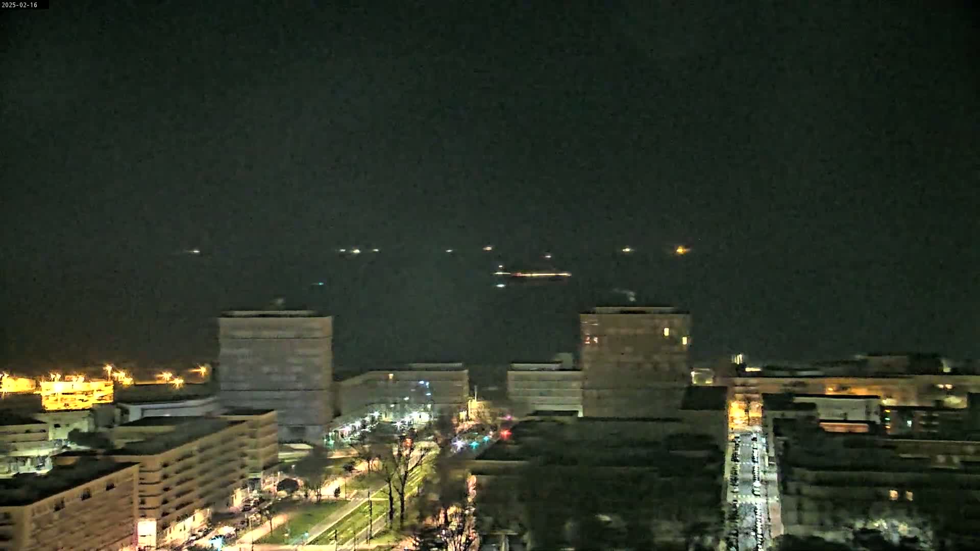 A nighttime view of a city skyline with several ships visible on the water in the distance under a dark sky.