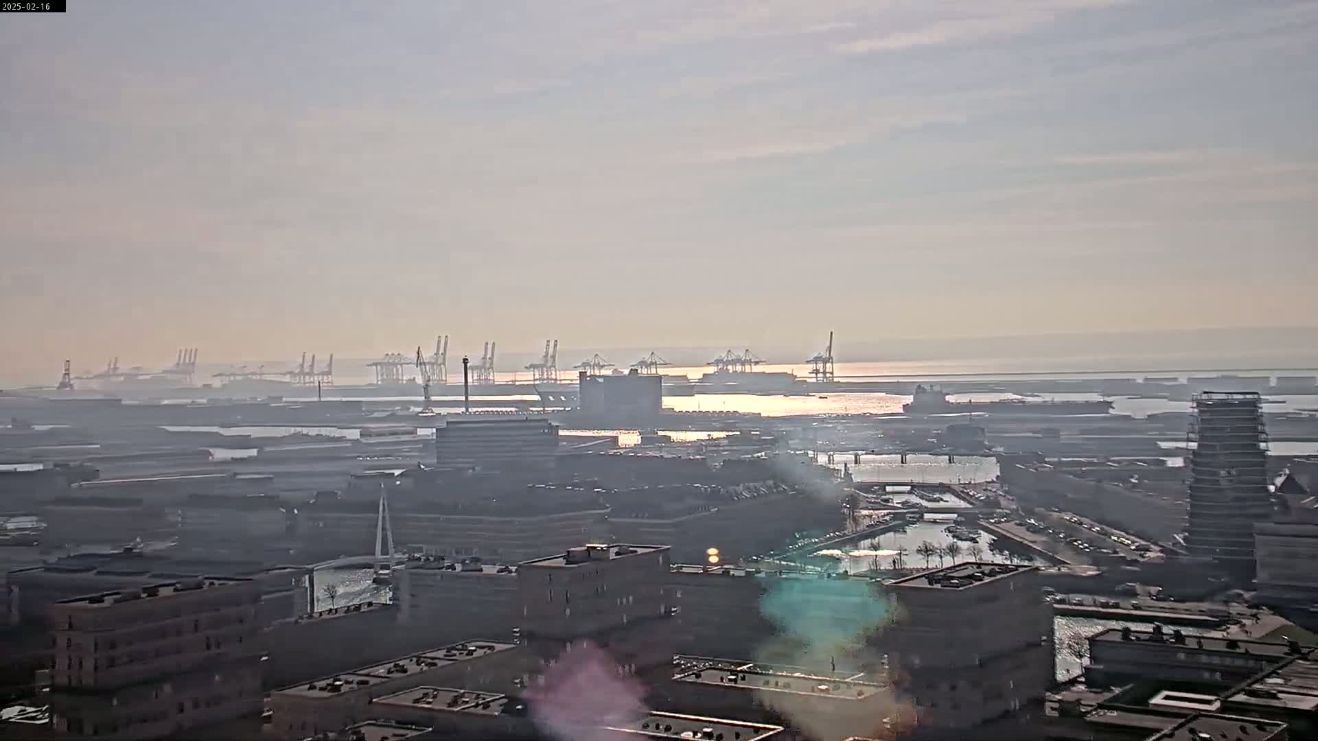 A hazy sunrise illuminates a sprawling industrial port area with numerous cranes and buildings, overlooking a calm body of water.