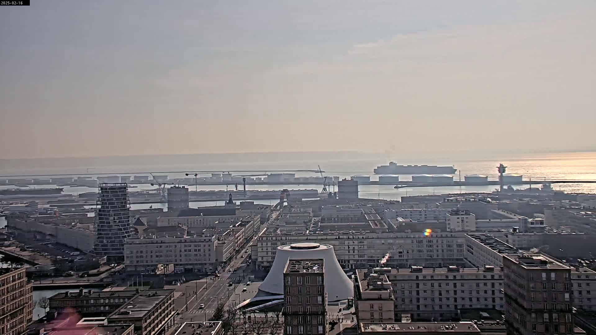 A hazy, sunny day reveals a port city with numerous buildings, a large container ship in the harbor, and a partially constructed skyscraper near the water.