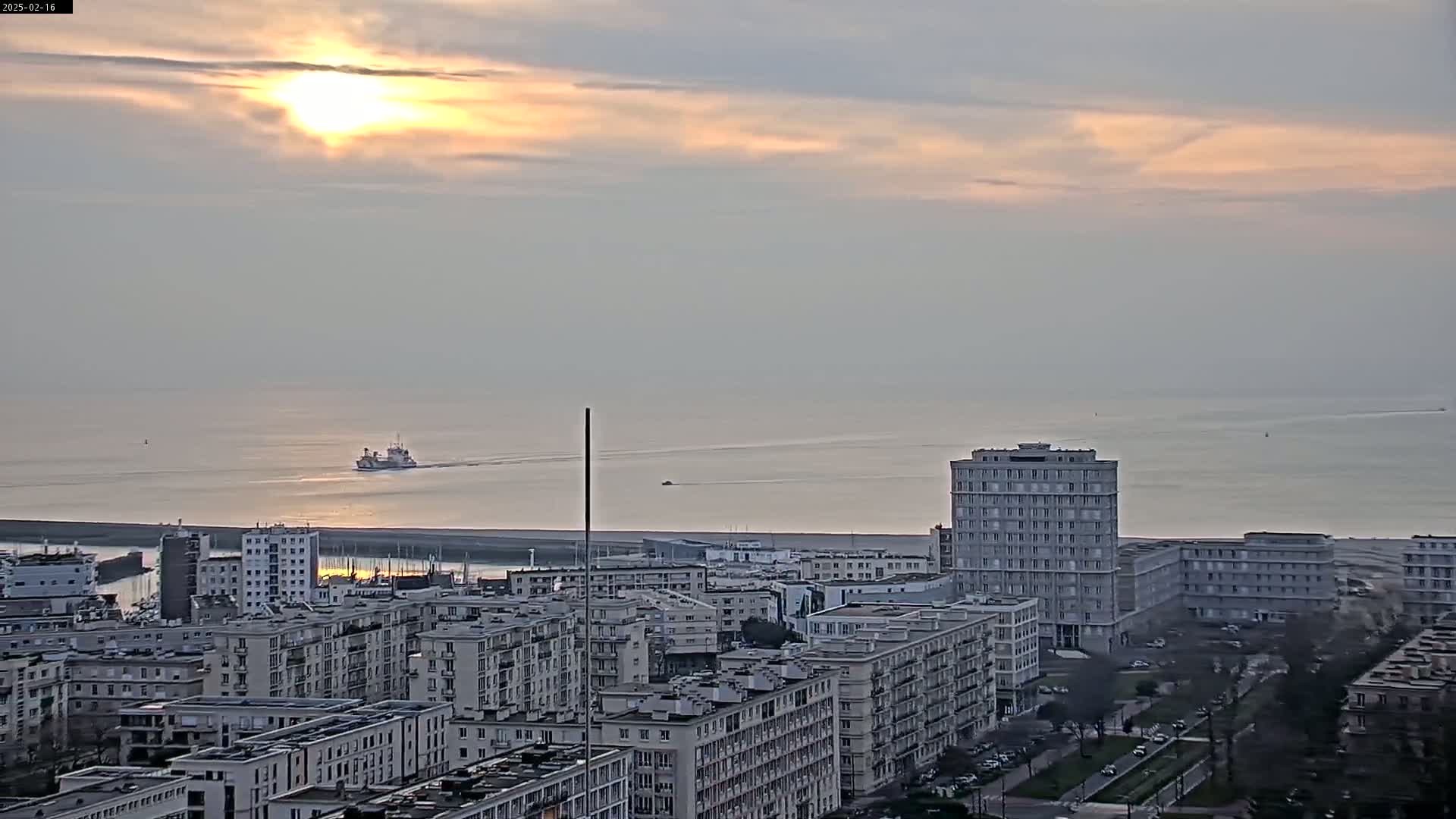 A city skyline with numerous apartment buildings is situated near a calm body of water where a ferry is visible, under a partly cloudy sunset sky.