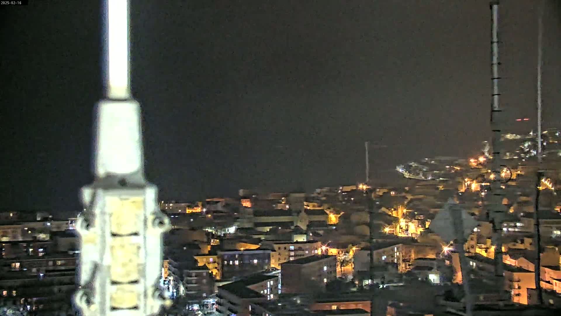 A nighttime, dark, and somewhat hazy view of a coastal town illuminated by scattered lights, seen from behind a tall metal structure.