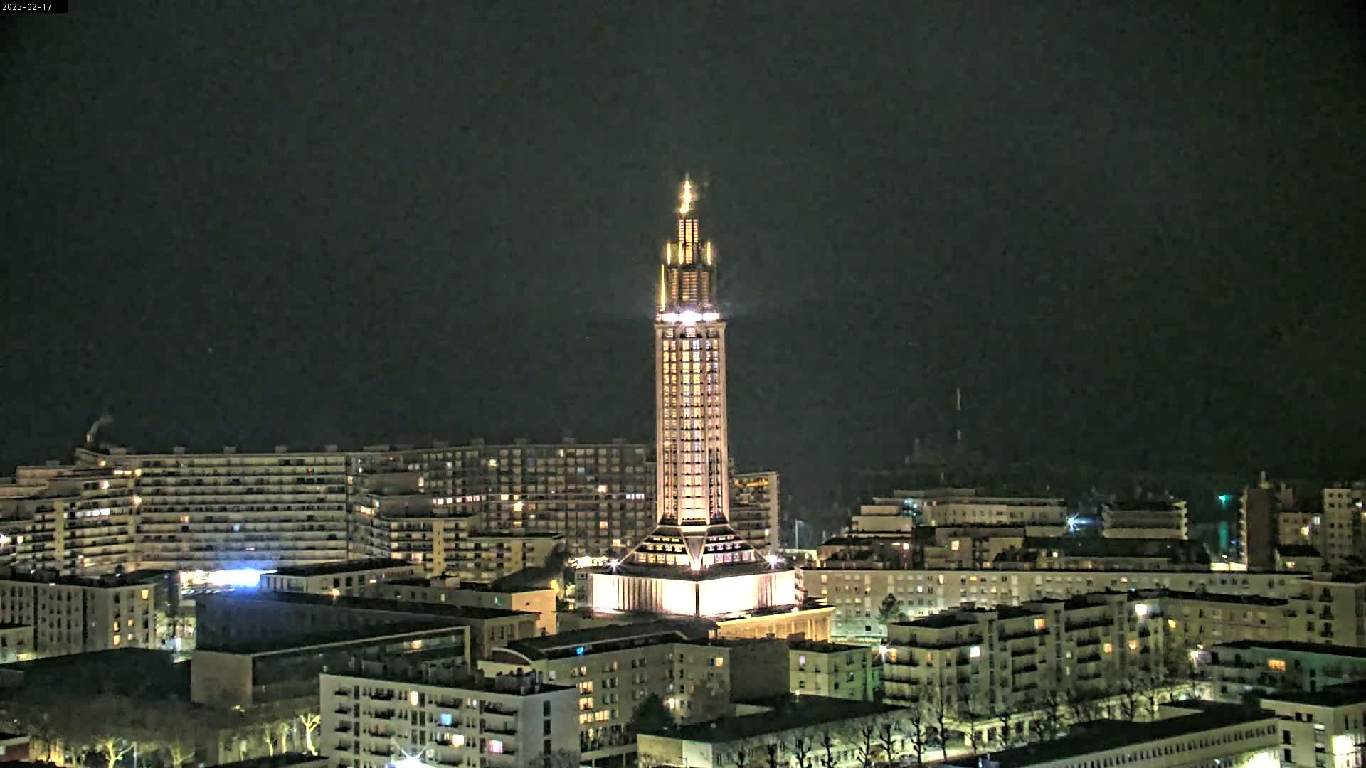A tall, illuminated tower stands in the center of a city at night under a dark sky.