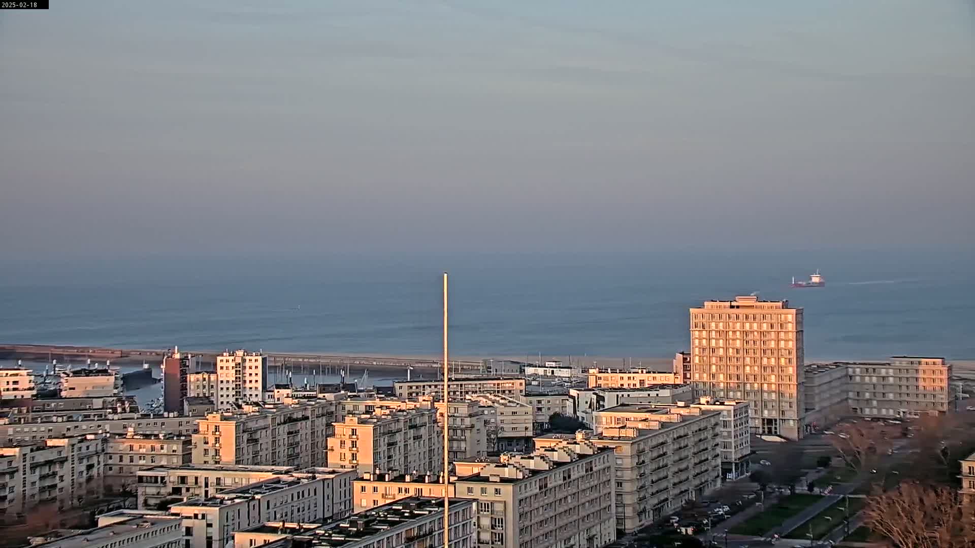 A coastal city with numerous apartment buildings is seen from an elevated viewpoint on a calm, clear evening, overlooking a tranquil sea with a single distant ship.