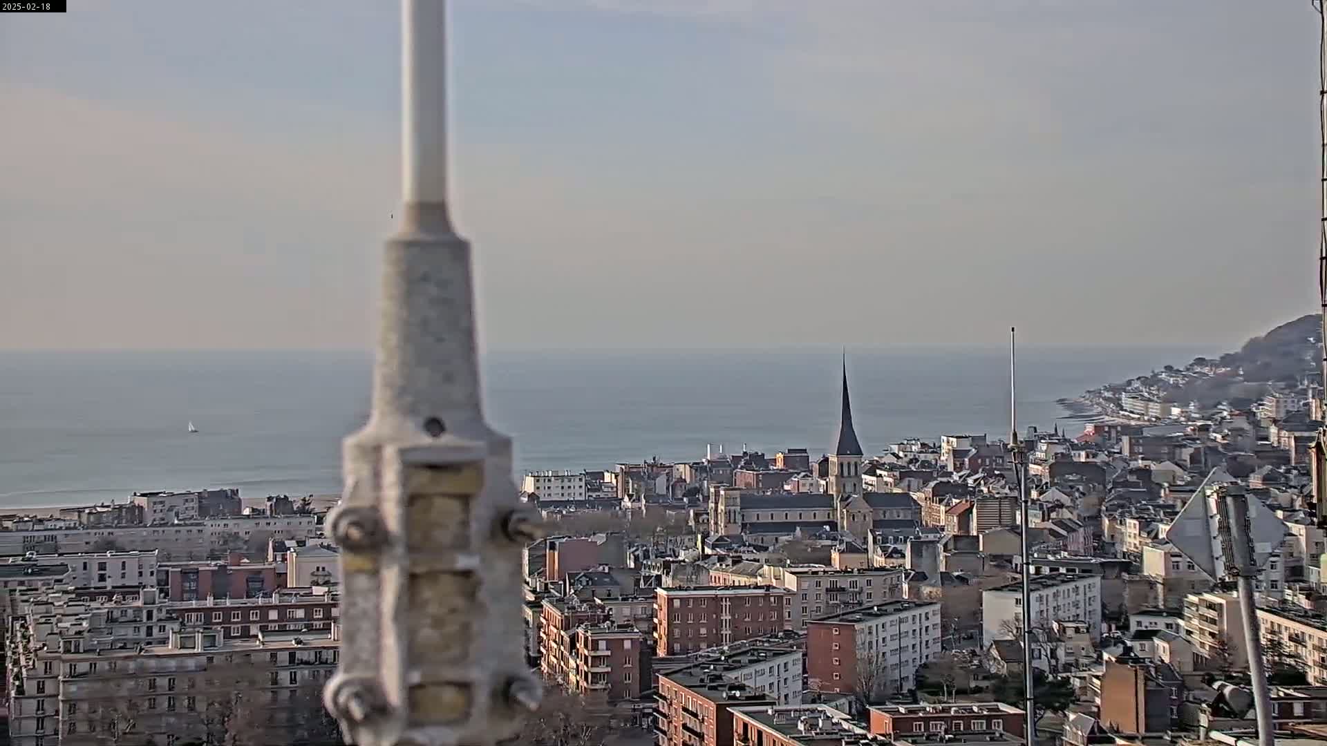 A coastal city with a church steeple is visible from a high vantage point on a hazy day, with a calm sea in the background.