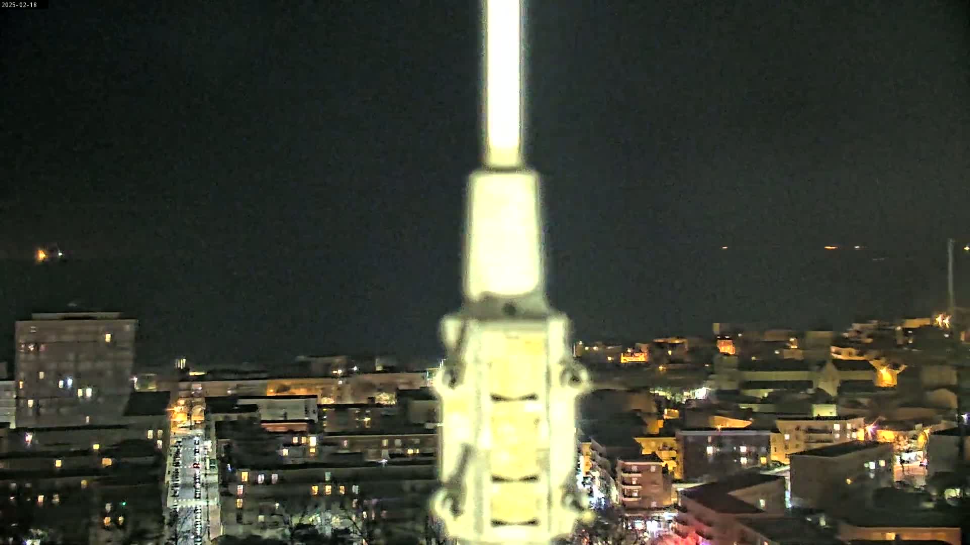 A nighttime view of a city illuminated by streetlights, with a tall, slender object in the center of the frame, and a dark, clear sky.