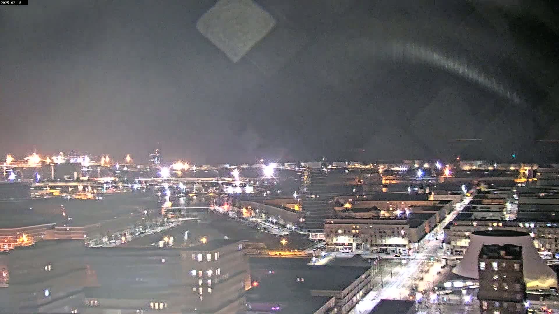 A nighttime, aerial view of a city with a harbor showing lights from buildings and vehicles under a dark, mostly clear sky.
