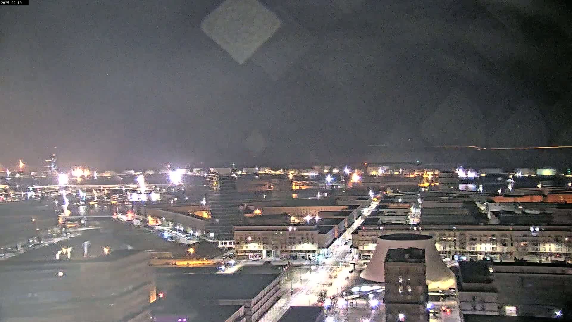 A nighttime, high-angle view of a city's illuminated streets and buildings, extending to a dark waterfront.