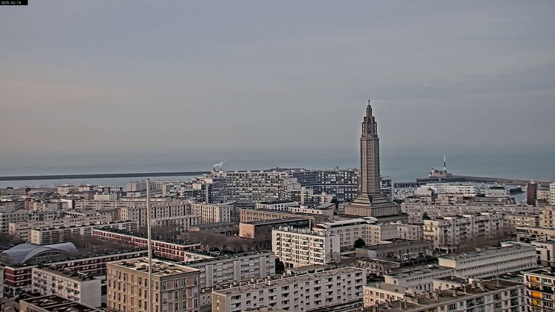 A coastal city with numerous apartment buildings and a tall tower is visible under a hazy sky, with calm ocean water in the background.
