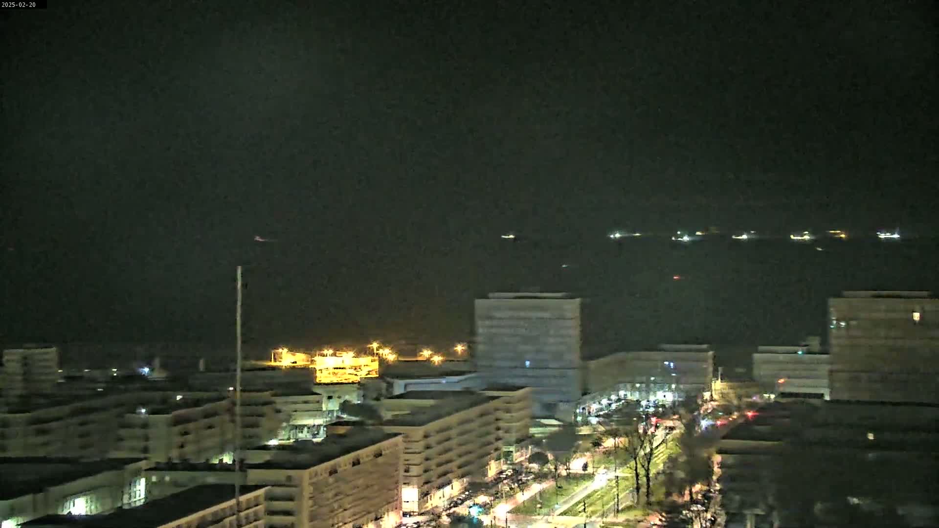 A nighttime cityscape with several buildings illuminated, overlooking a dark body of water where lights from ships are visible in the distance under a clear sky.