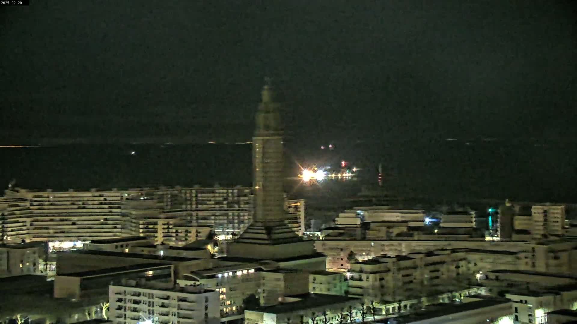 A tall tower stands in a city at night, overlooking a dark body of water under a clear, dark sky.