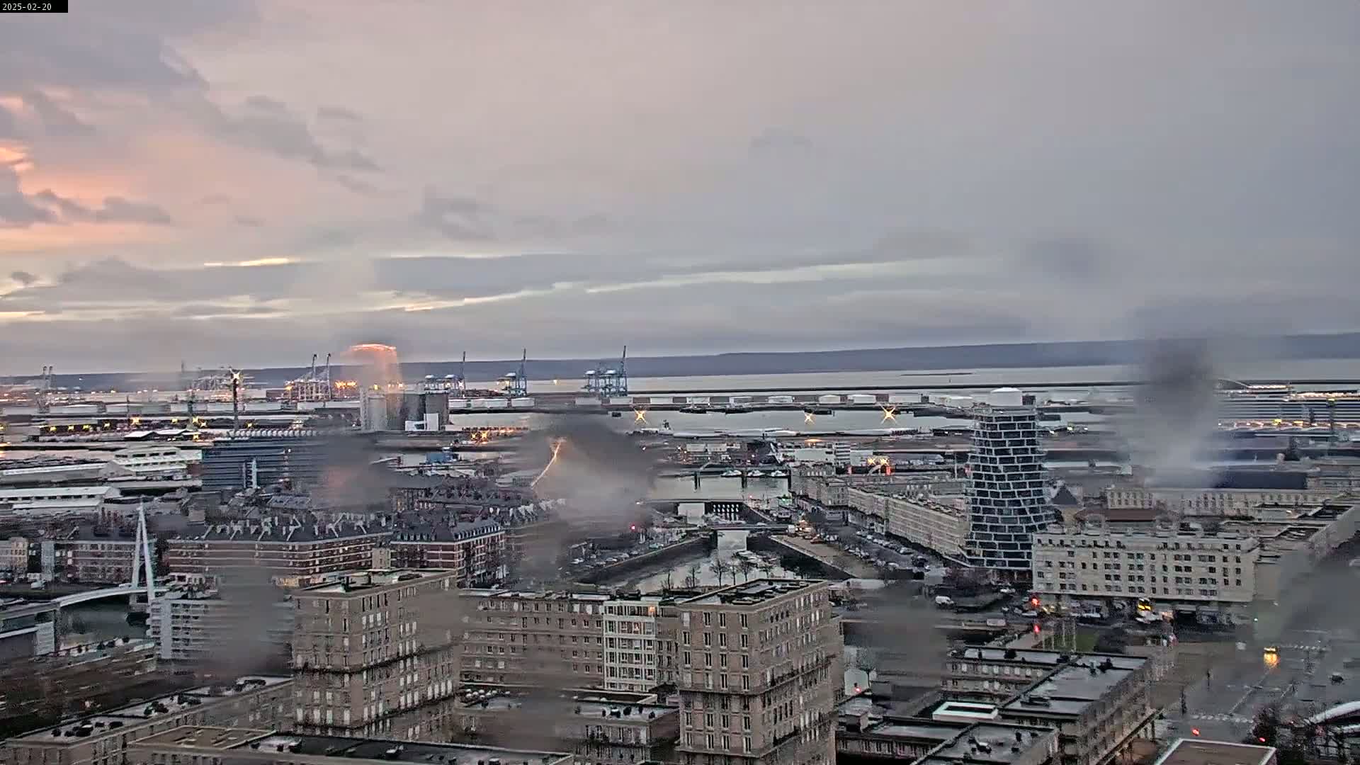 A large city with a harbor is partially obscured by smoke from a fire under an overcast sky.