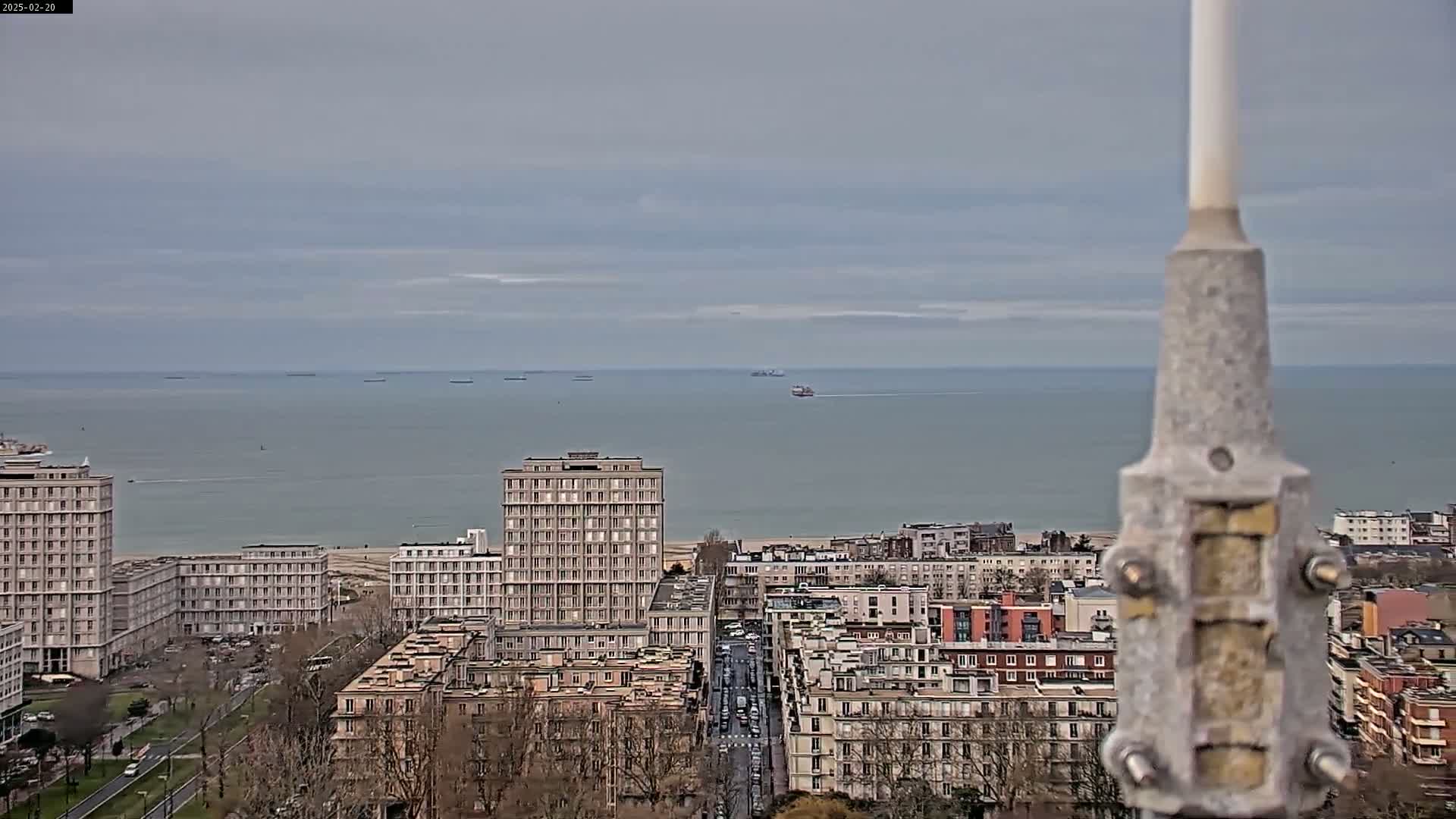 A city overlooking a calm, gray sea under a cloudy sky, with several tall buildings and a street visible in the foreground.
