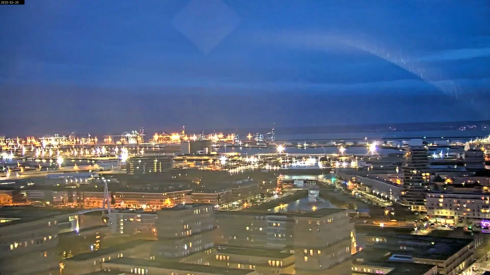 A nighttime aerial view of a city harbor with many lights illuminating buildings and industrial areas under a clear, dark blue sky.
