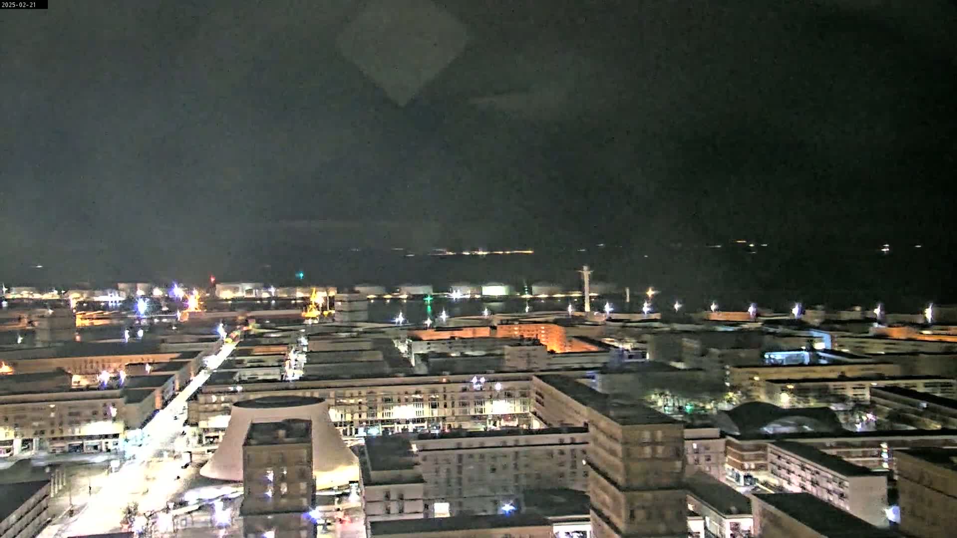 A nighttime aerial view of a city harbor with numerous buildings illuminated, under a dark, mostly cloudy sky.
