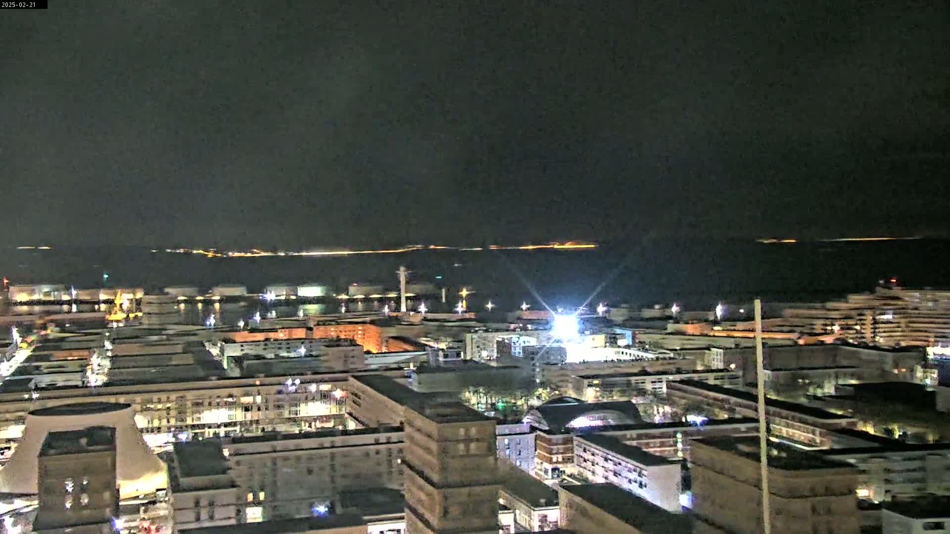 A nighttime aerial view of a city harbor, with numerous lights illuminating buildings and the waterfront, under a clear, dark sky.