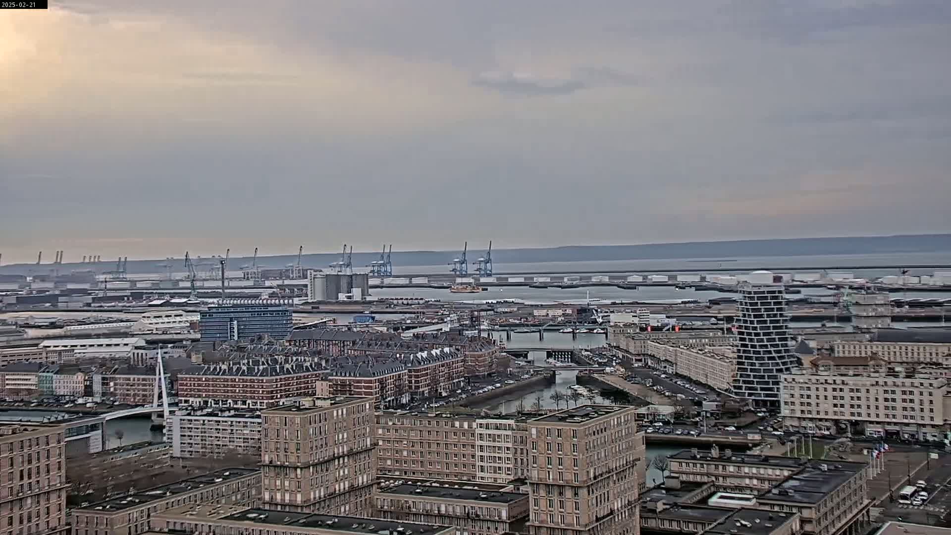 A panoramic view of a city's port area on an overcast day, showing numerous buildings, a canal, and industrial docks with cranes extending into a calm body of water.