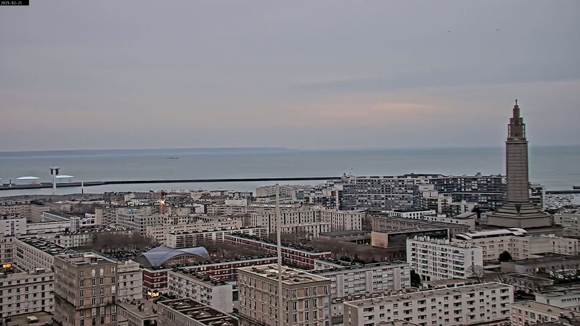 A coastal city with numerous buildings and a tall tower is visible under an overcast sky, with a calm body of water in the background.