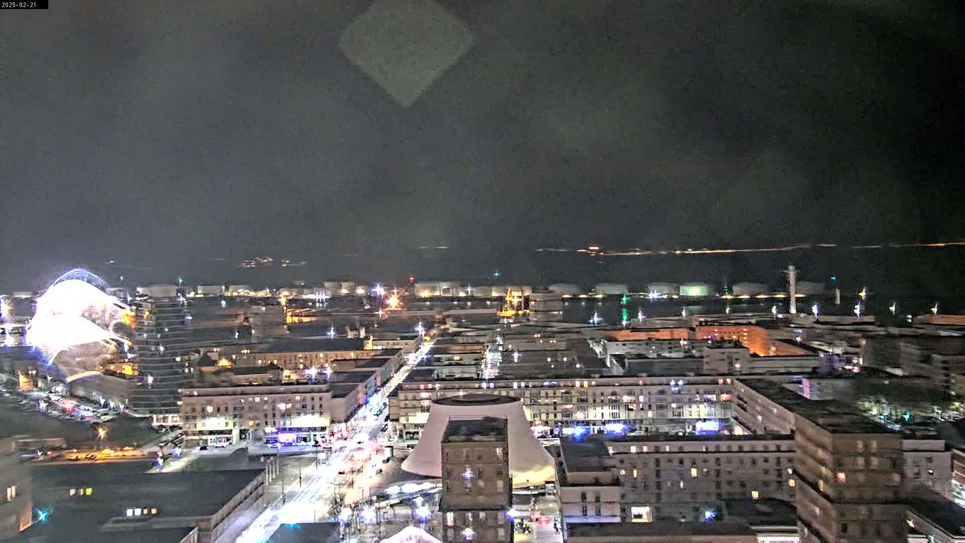 A nighttime aerial view of a city with a body of water in the distance, showing illuminated buildings and streets, with a faint, possibly hazy sky.