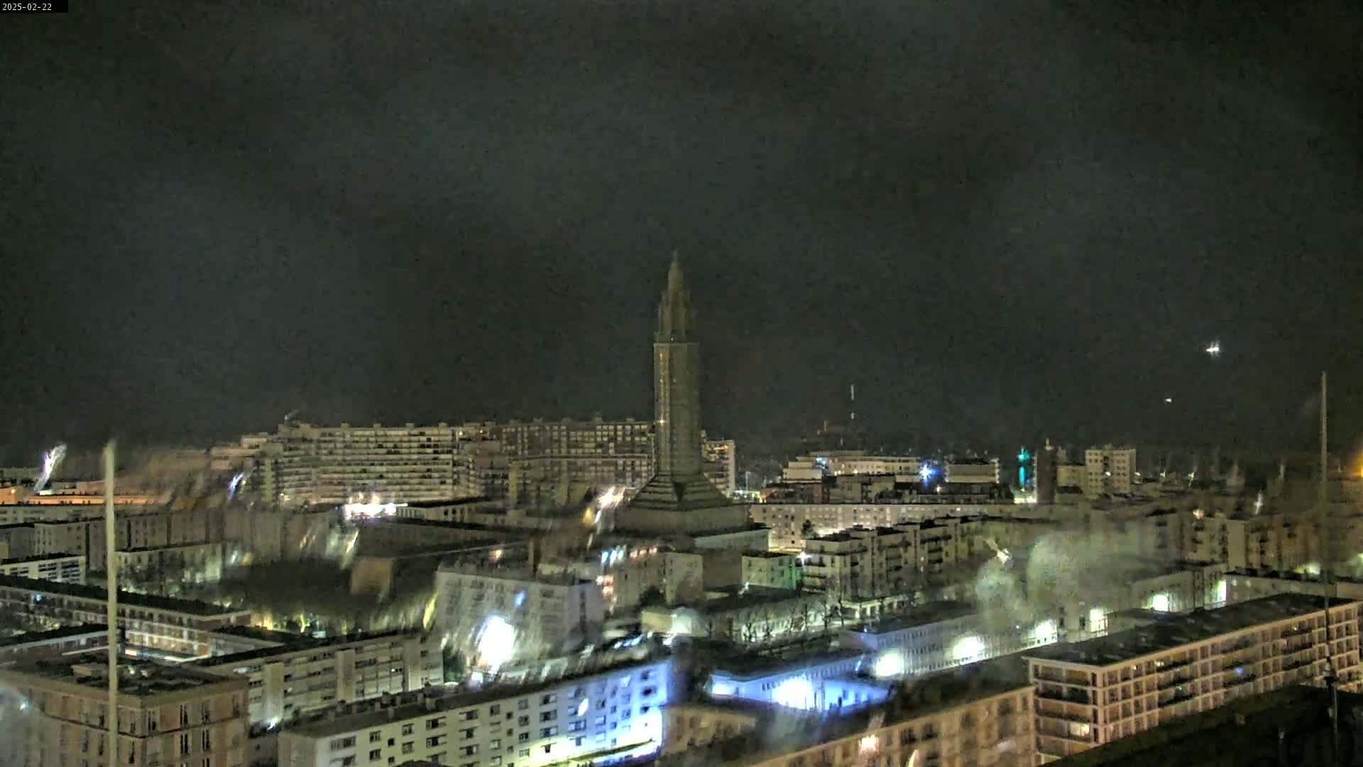 A nighttime aerial view of a city with a tall tower in the center, under a dark, cloudy sky.