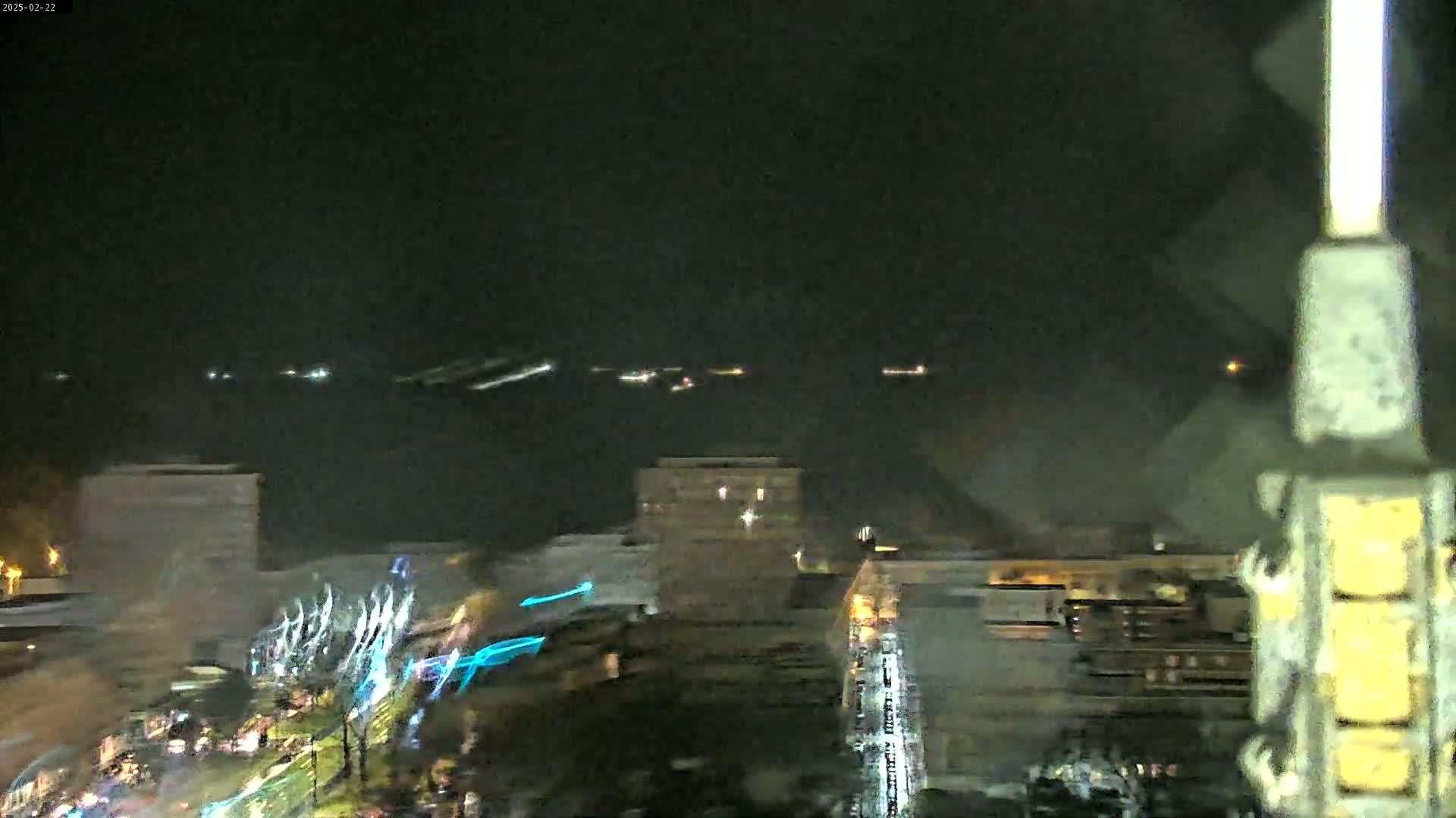 A nighttime view of city buildings and a body of water with faint lights in the distance, under a dark, possibly overcast sky.