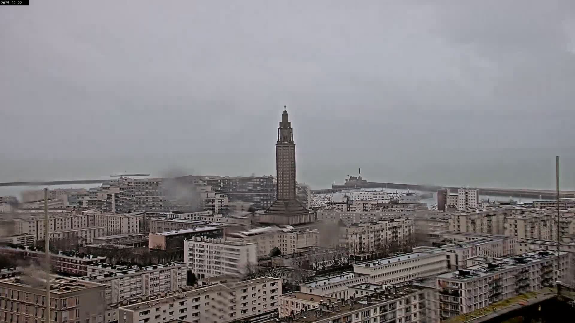 A tall, slender tower stands prominently in the center of a city overlooking a gray, overcast ocean.