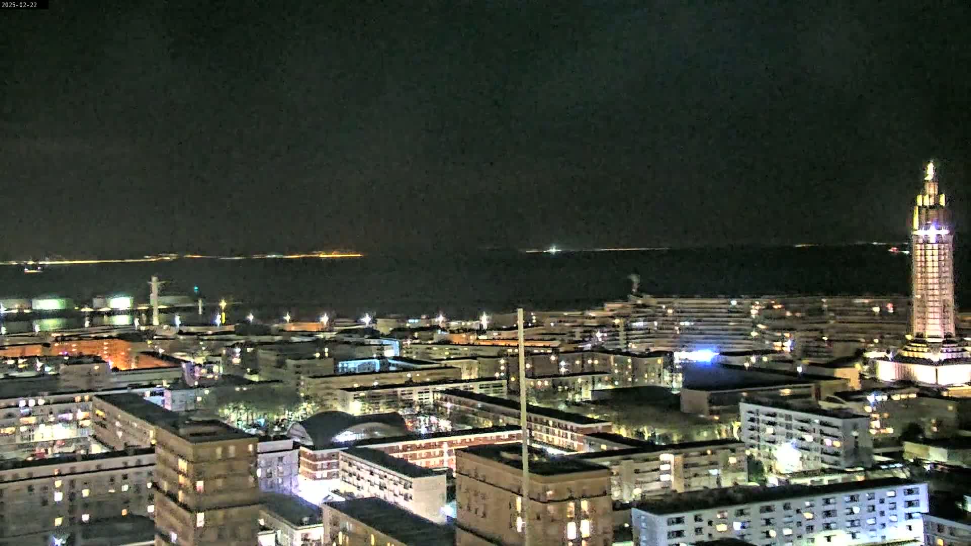 A nighttime aerial view of a city with a tall tower at the right, overlooking a calm body of water under a dark sky.