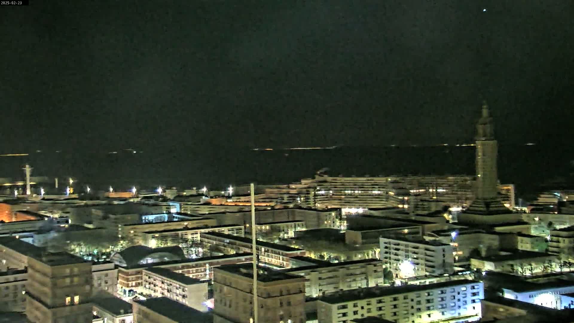 A nighttime aerial view of a city by a body of water under a clear, dark sky shows numerous buildings illuminated with lights.